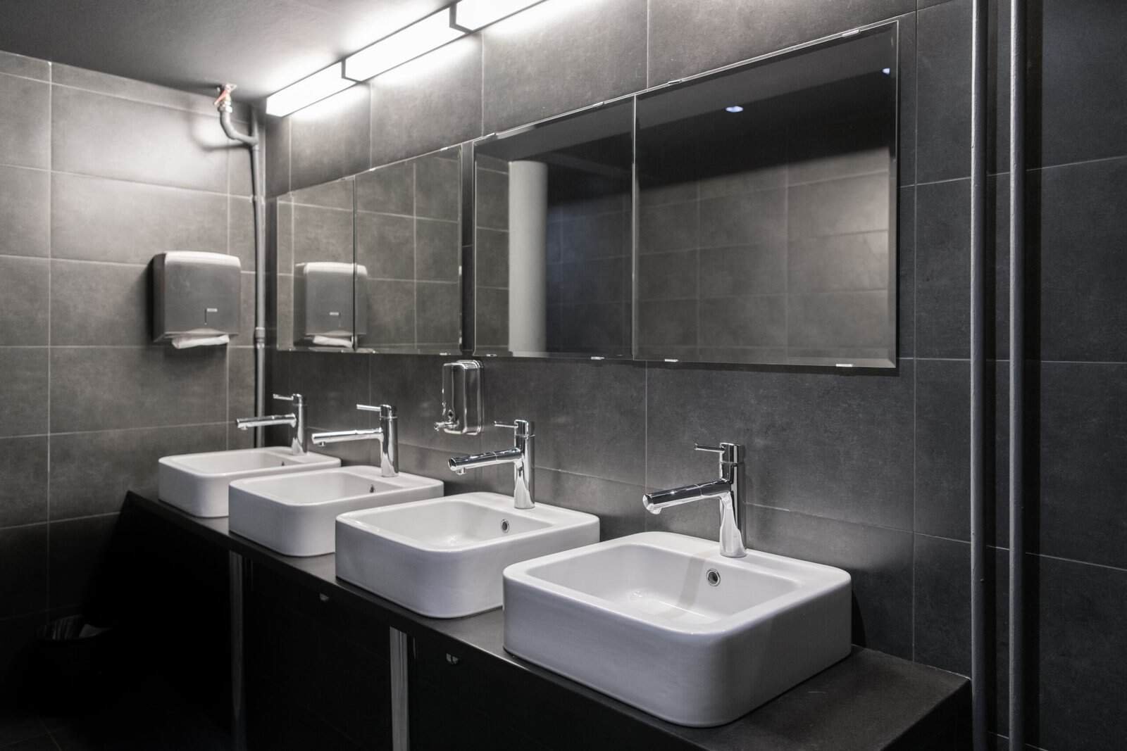 Modern public restroom with four white sinks and large mirror on dark gray tiled wall.