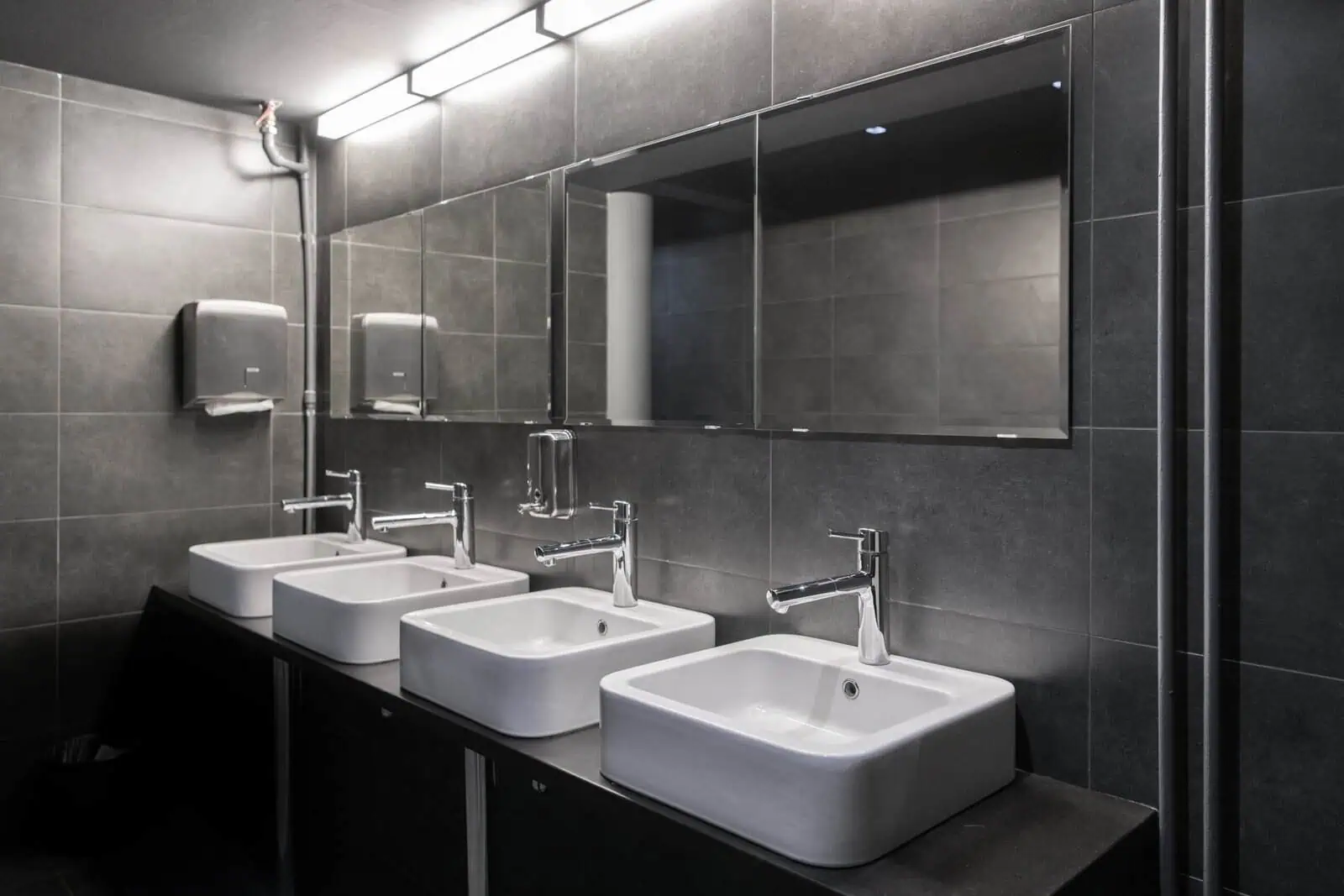 Modern public restroom with four white sinks and large mirror on dark gray tiled wall.